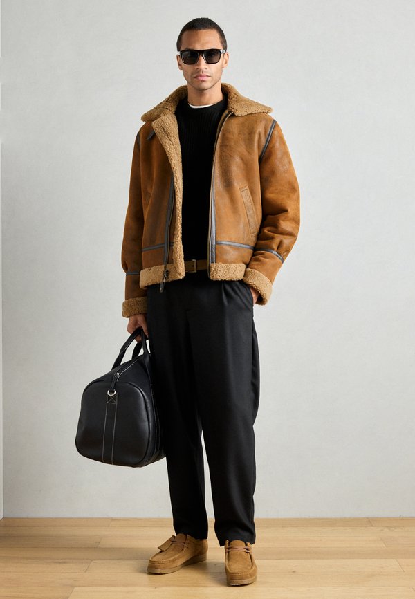 Jaqueta de shearling castanha com um colarinho de pele, detalhes cinzentos, suéter preto texturizado, calças pretas largas e sapatos de camurça castanhos; segurando uma bolsa preta.