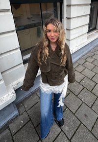 Jeune femme aux cheveux blonds ondulés portant une veste vert olive, un haut en dentelle blanche, un jean large bleu et des chaussures noires, debout sur le trottoir près d'un bâtiment blanc.