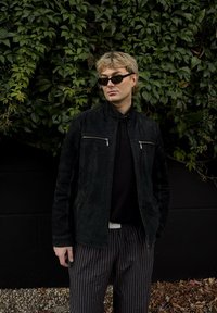 Chaqueta de ante negra con cremalleras plateadas, camiseta polo negra y pantalones anchos a rayas. Contrasta con un fondo de follaje verde.