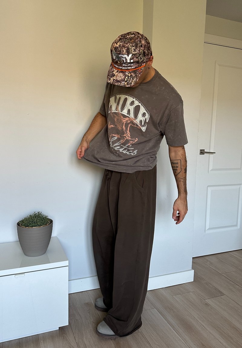 T-shirt graphique Nike marron avec un look usé, associé à un pantalon marron ample. Une casquette camouflage vient compléter la tenue.