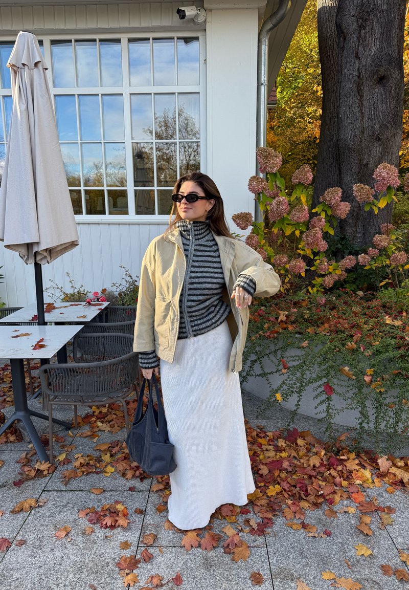Jupe longue crème, col roulé rayé gris et noir, veste beige. Lunettes de soleil noires et sac à main, entourée de feuilles et de fleurs d'automne.