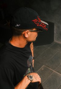 Man wearing a black cap with a red flame design, a black graphic t-shirt, glasses, and a gold and silver wristwatch, looking to the right.