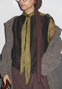 Tenue superposée comprenant un chemisier marron semi-transparent avec un nœud jaune, un manteau gris texturé, et une écharpe bordeaux. Les accessoires incluent des boucles d'oreilles rondes.