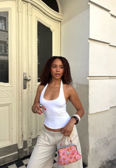 Mujer con cabello rizado, vestida con un top halter blanco y pantalones beige, sostiene un bolso floral rosa, parada junto a una puerta amarillo pálido en una calle de la ciudad.