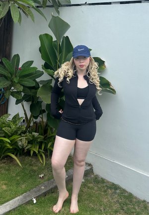 Chaqueta negra con cremallera y diseño recortado, combinada con un sujetador deportivo negro y pantalones cortos. Se lleva con una gorra de béisbol azul marino, descalzo cerca de vegetación.