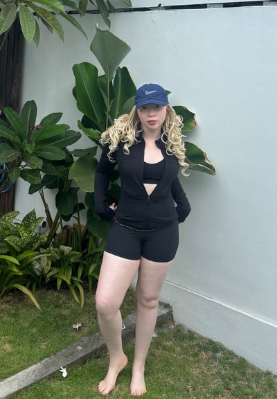 Chaqueta negra con cremallera y diseño recortado, combinada con un sujetador deportivo negro y pantalones cortos. Se lleva con una gorra de béisbol azul marino, descalzo cerca de vegetación.