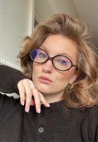 Femme aux cheveux bouclés châtain clair portant des lunettes et un pull sombre, reposant son menton sur sa main, à l'intérieur avec un arrière-plan neutre.