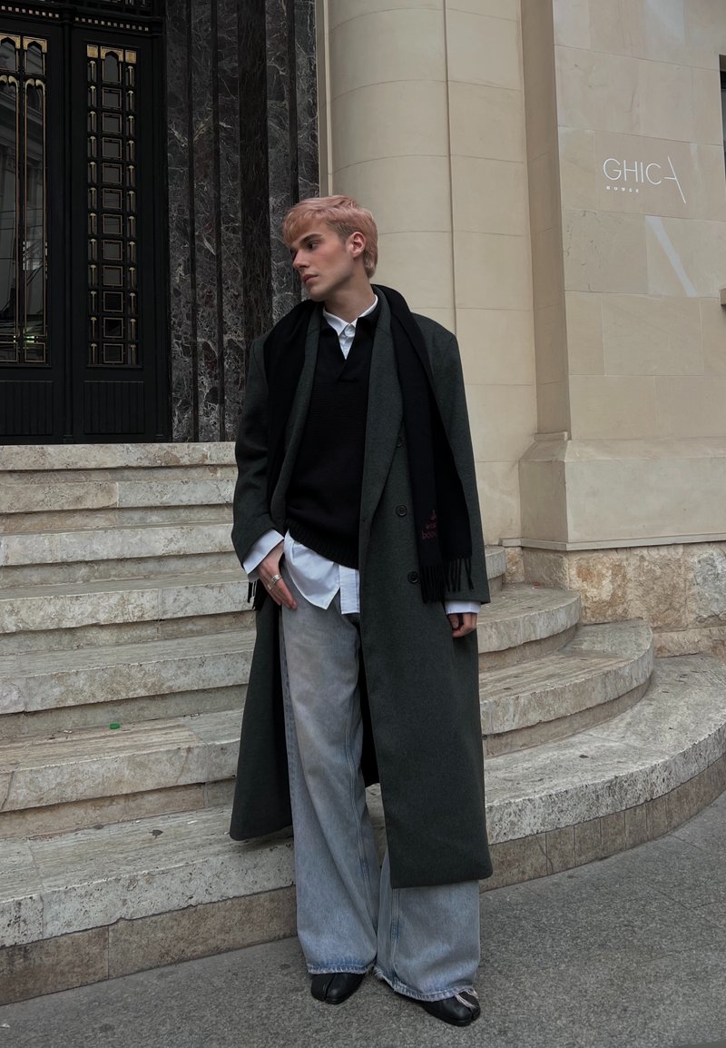 Long manteau gris sur pull noir, chemise blanche et jean large bleu clair. Écharpe noire drapée, debout sur des marches en pierre dans un cadre urbain.
