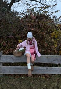 Lichtroze oversized trui, witte korte broek met kanten rand, roze en bordeauxrood patroon sjaal, crèmekleurige kniekousen, cognackleurige laarzen, rieten mand met groenblijvende planten.