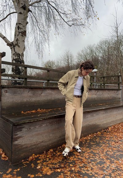 Chaqueta beige sobre una camisa blanca, combinada con pantalones beige holgados y zapatos con patrón en blanco y negro. Banco de madera y hojas de otoño de fondo.