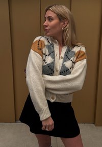 Λευκό πλεκτό κασκόλ με πολύχρωμο διαμαντένιο σχέδιο σε γκρι, πορτοκαλί και μαύρο. Διαθέτει κουμπιά και ριμπ μανσέτες, φορεμένο πάνω από μια μαύρη φούστα.