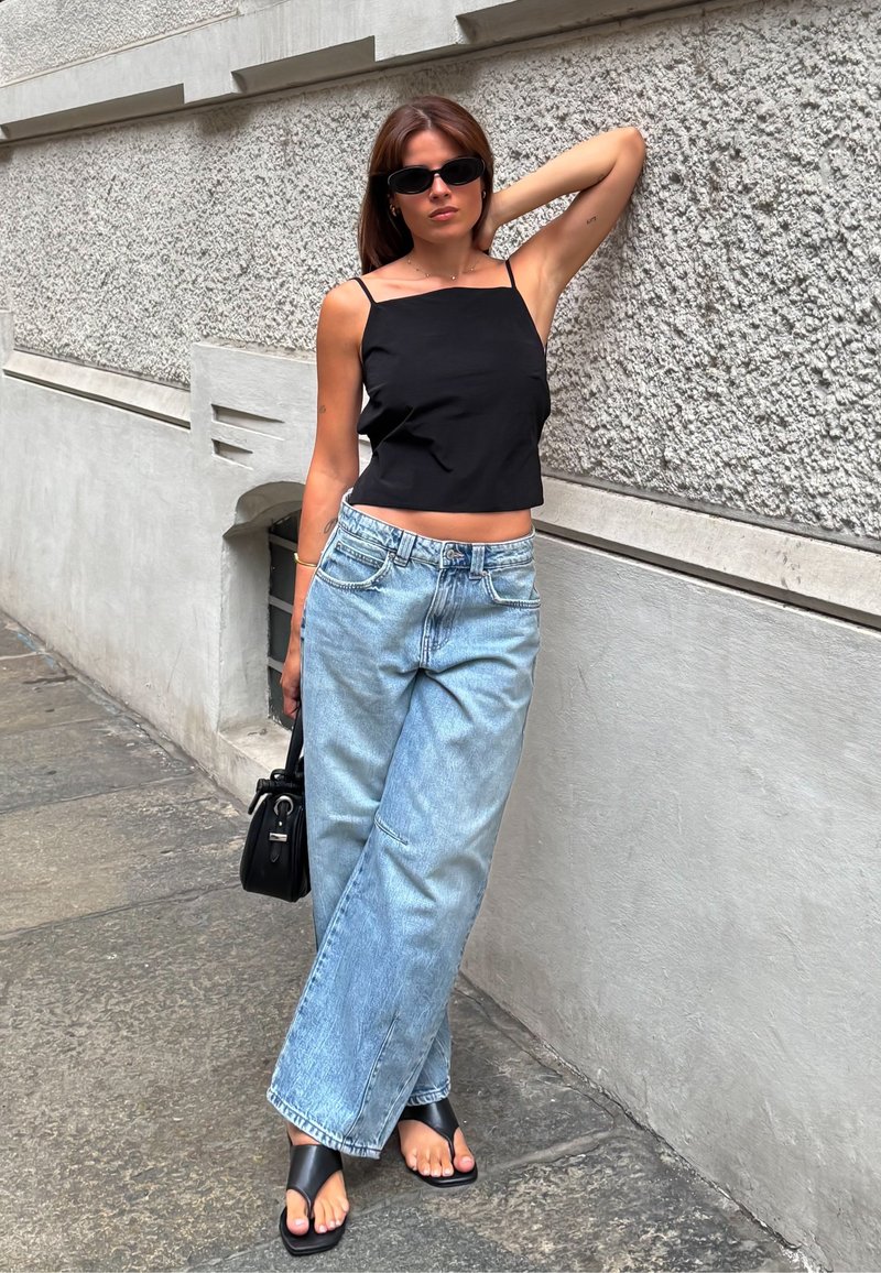 Top cropped nero, jeans blue chiari a gamba larga, sandali neri piatti. Gli accessori includono una piccola borsa nera e occhiali da sole. Sfondo con parete testurizzata.