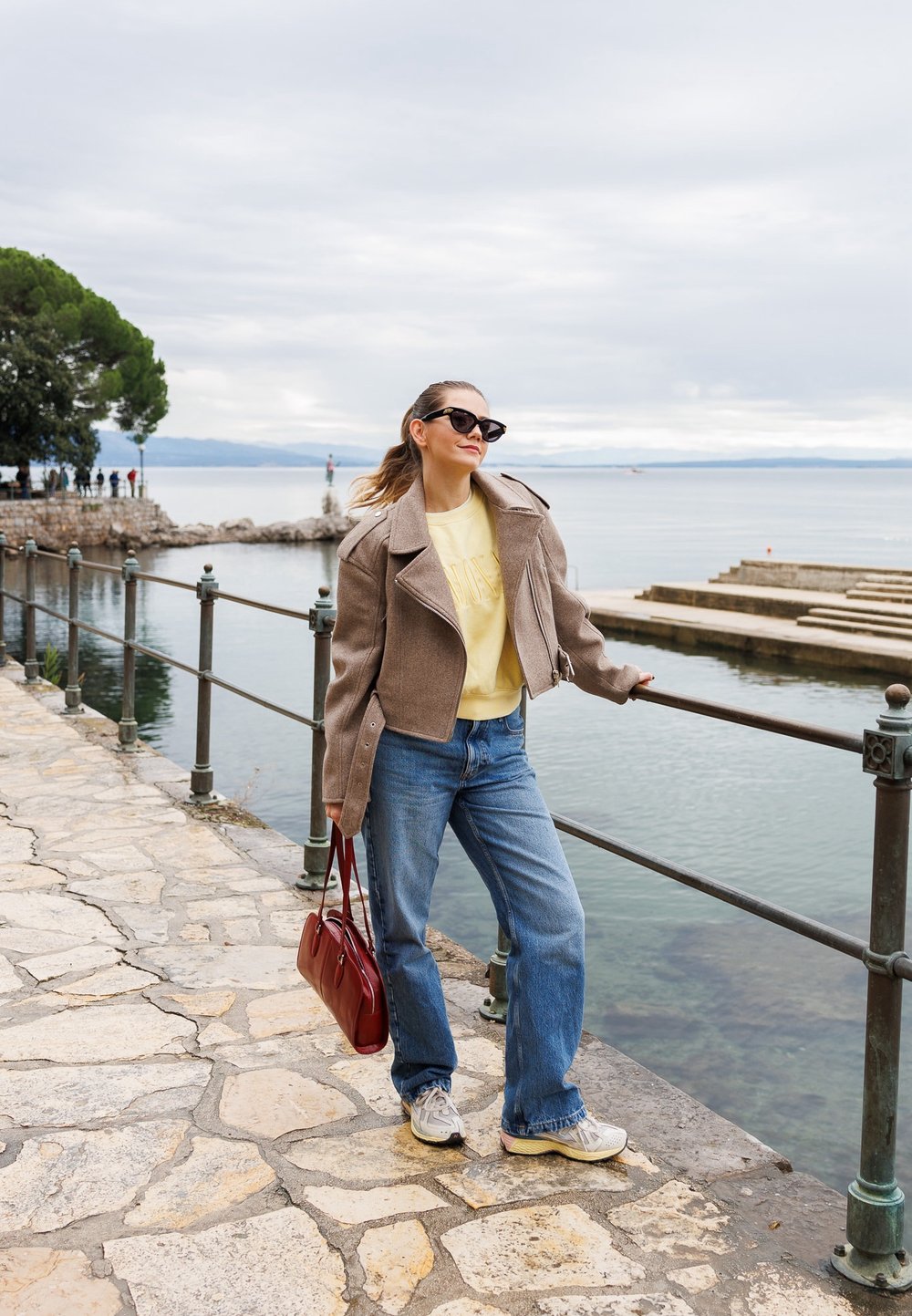 Veste de moto en laine marron, sweat-shirt jaune, jeans bleus et baskets grises. Tient un sac à main rouge. Chemin en pierre à côté d'une promenade au bord de l'eau.