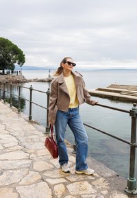 Veste de moto en laine marron, sweat-shirt jaune, jeans bleus et baskets grises. Tient un sac à main rouge. Chemin en pierre à côté d'une promenade au bord de l'eau.
