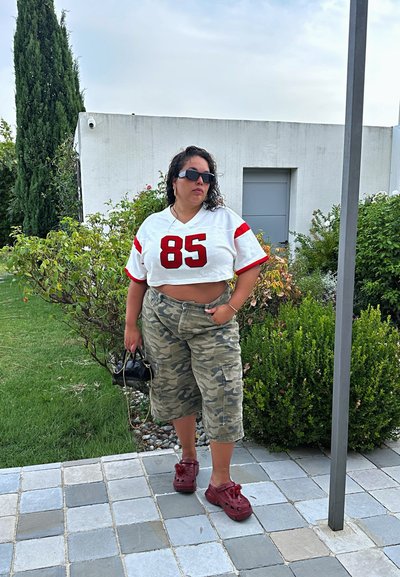 Jersey blanco recortado con acentos rojos y "85", pantalones cortos de camuflaje, zapatos de madera rojos y bolso negro, en un área verde con arbustos.