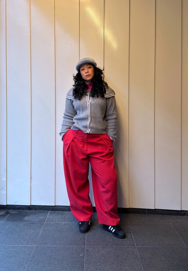 Kvinde med langt krøllet hår iført en grå strikket cardigan, matchende baret, løse røde bukser og sorte sneakers, stående op ad en beige væg.