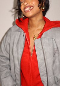 Veste matelassée grise avec une capuche rouge portée sur un pull zippé rouge texturé, dotée d'une fermeture éclair et d'un collier pendentif.