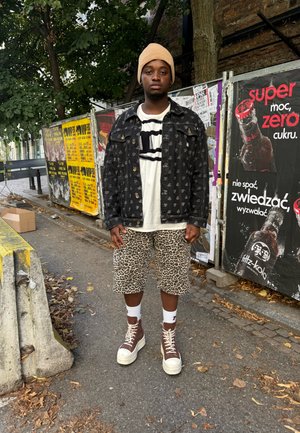 Chaqueta de mezclilla negra con logos, camiseta gráfica blanca, pantalones cortos de estampado de leopardo, zapatillas altas marrones y gorro de punto beige. Entorno urbano.