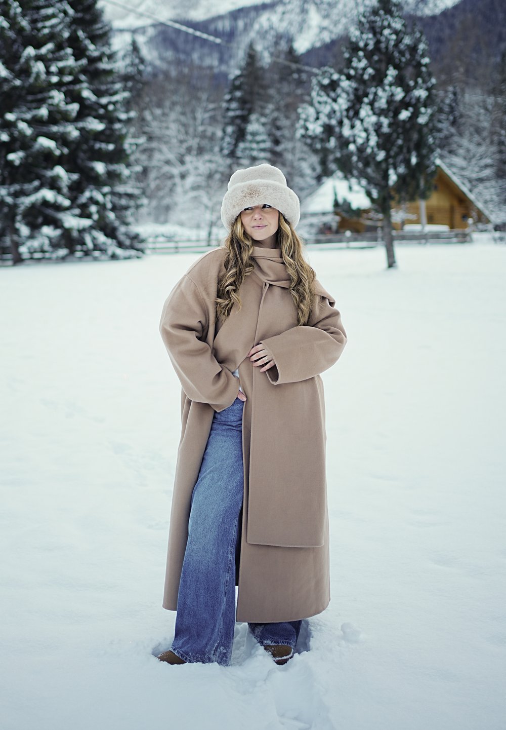 Femme en manteau beige, chapeau beige et jean, debout dans un champ enneigé avec des arbres et une cabine en arrière-plan pendant l'hiver.