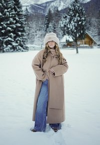 Femme en manteau beige, chapeau beige et jean, debout dans un champ enneigé avec des arbres et une cabine en arrière-plan pendant l'hiver.