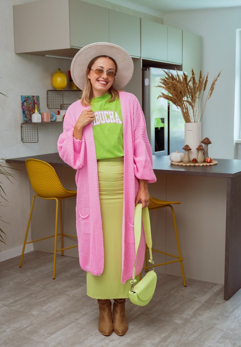 Lunga cardigan rosa di maglia sopra una camicia verde, gonna gialla e stivali marroni. Tiene una borsa verde piccola. Sfondo di una cucina moderna.