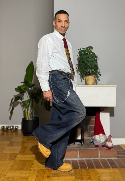 Hombre con camisa blanca, corbata estampada, jeans anchos y botas amarillas posando en el interior junto a una planta y una chimenea con decoraciones de gnomos.