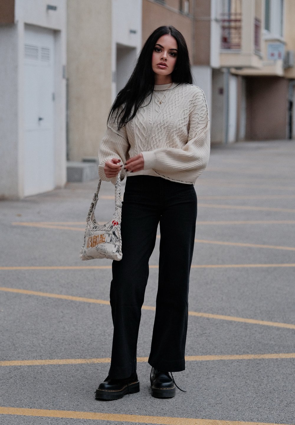 Pullover chunky in crema a trecce, pantaloni neri a palazzo, scarpe platform nere e una borsa a spalla con un design grafico.