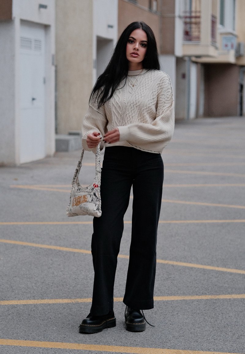 Chunky cream cable-knit sweater, black flared trousers, black platform shoes, and a patterned shoulder bag featuring a graphic design.