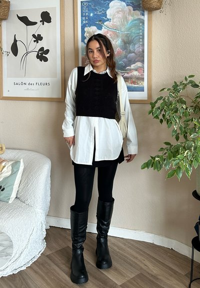 Chaleco de punto negro sobre una camisa blanca de botones, combinado con leggings negros y botas negras hasta la rodilla; ambiente interior con plantas.