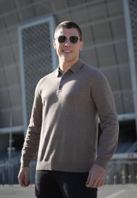 Light brown knit polo shirt with long sleeves, three buttons at the collar, worn with black pants. Individual wearing black sunglasses.