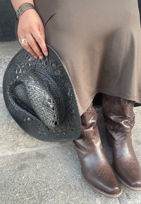 Μαύρο πλεγμένο καπέλο από ψάθα κρατημένο σε ένα χέρι, καφέ φόρεμα, καφέ καουμπόικες μπότες με διακοσμητική ραφή και λεία υφή στο δέρμα της μπότες.