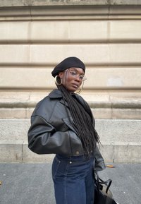 Zwarte leren oversized jas, high-waisted donkere spijkerbroek, zwarte baret, ronde bril, grote oorringen, staand tegen een textuurrijke muur.