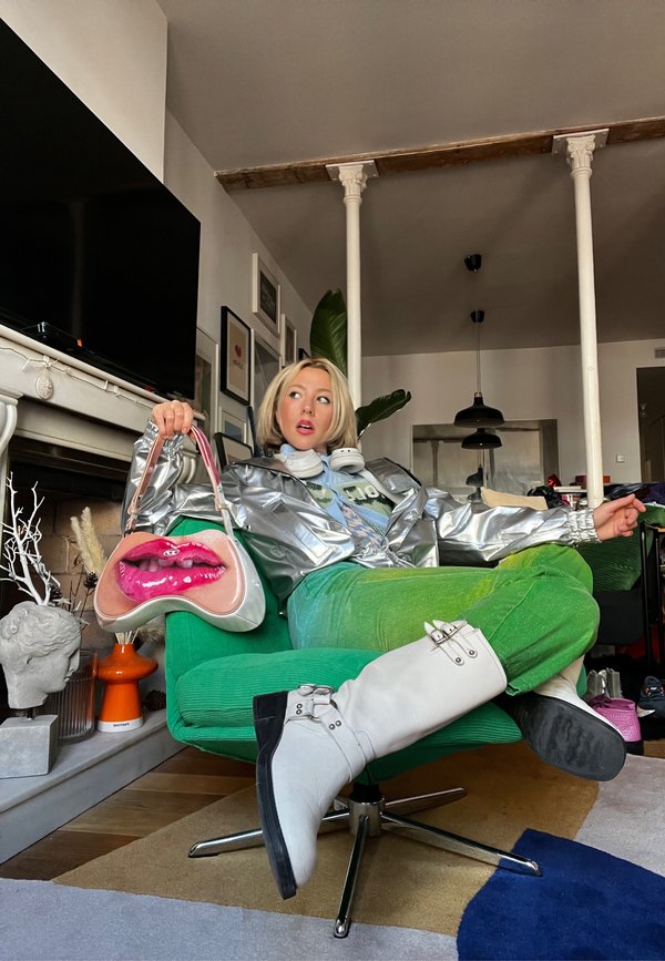 Giacca argentata metallica con zip, pantaloni verdi texture, stivali bianchi fino al ginocchio e borsa novità a forma di labbra. Sedia verde sullo sfondo.
