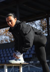 Impermeável desportivo preto com capuz, leggings justas pretas, a atar sapatilhas desportivas azul-claro, sentado num banco num ambiente de estádio.