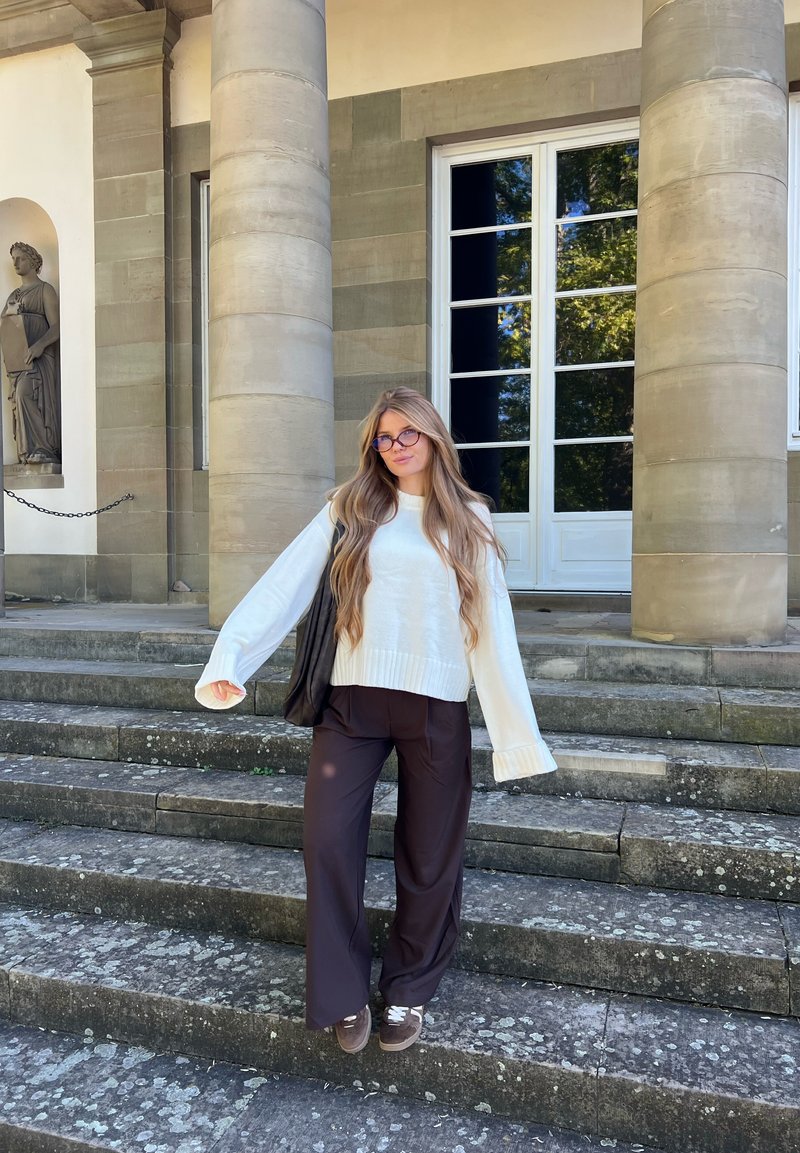 Jeune femme aux longs cheveux et portant des lunettes se tient sur des marches en pierre, portant un pull blanc, un pantalon sombre et portant un sac noir, devant des colonnes et des portes.