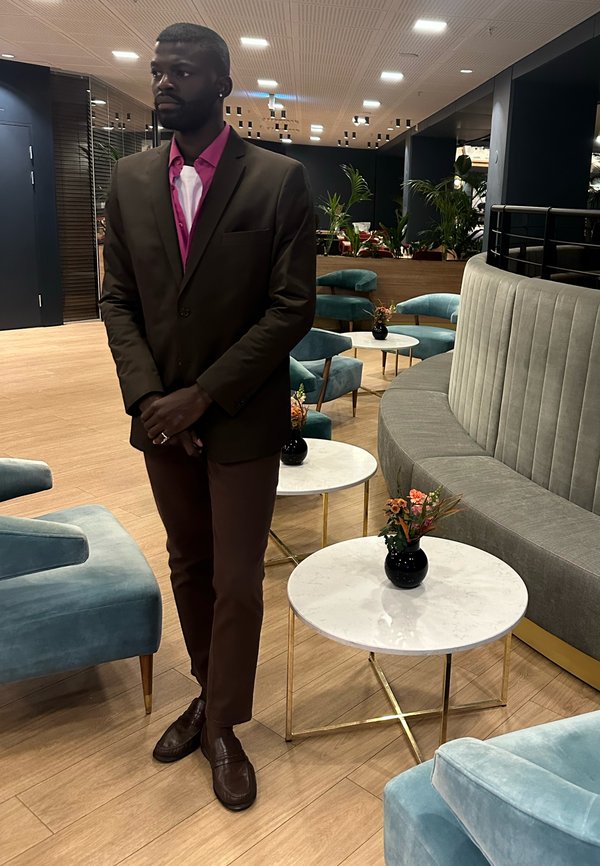 Bruin pak met een roze overhemd, staand naast lichte marmeren tafels en beklede zitplaatsen. Glanzende schoenen en een klein vaasje met bloemen in beeld.