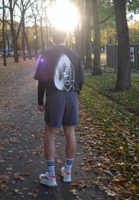 Czarna koszulka z abstrakcyjnym białym wzorem, szare szorty, białe sneakersy z różowymi akcentami, paskowane skarpetki, stojący na ścieżce pokrytej liśćmi.