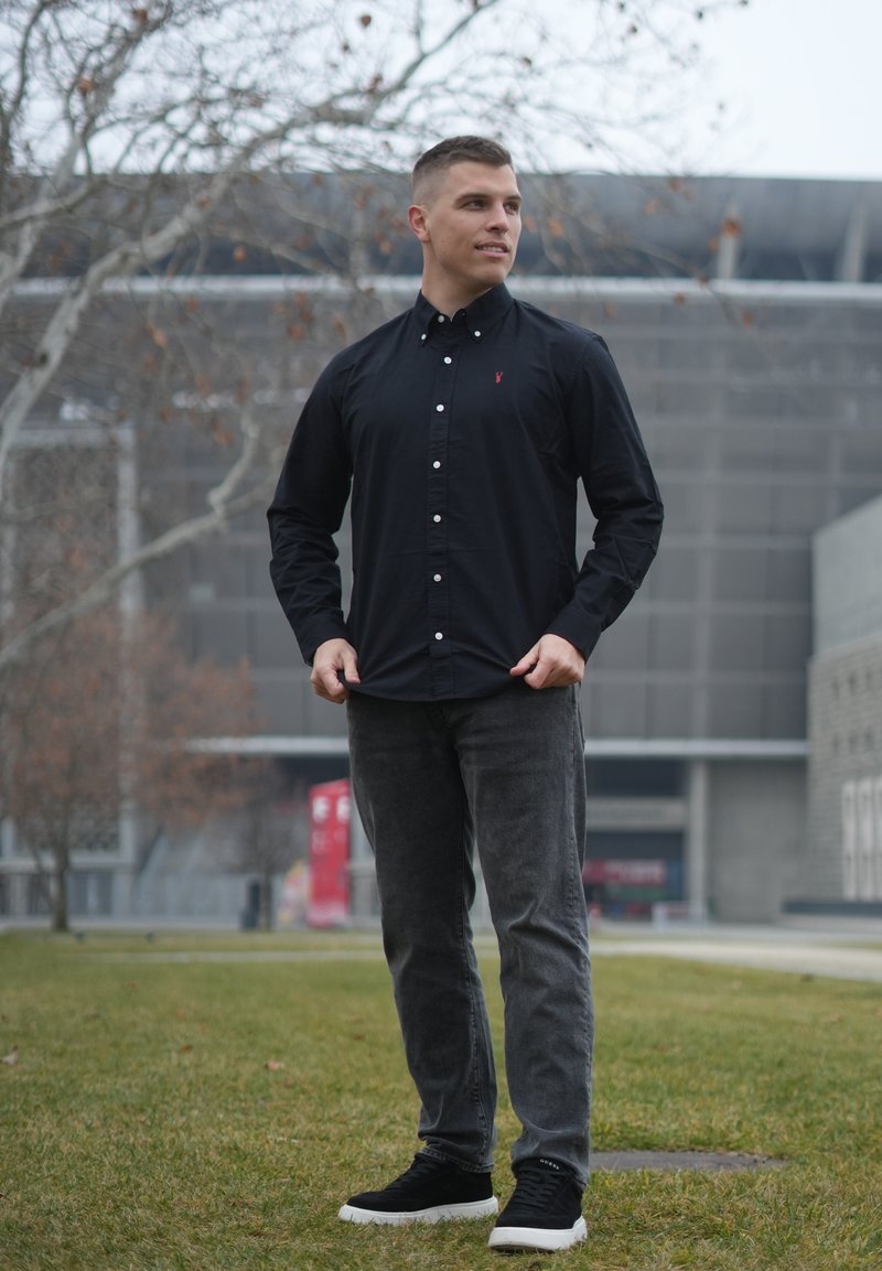 Camicia nera con bottoni, jeans grigi e scarpe da ginnastica nere. La camicia presenta un piccolo logo rosso, con una texture liscia e una vestibilità su misura. Ambientazione all'aperto.