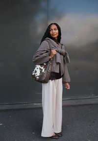 Vrouw met gevlochten haar, gekleed in een bruine trui en beige broek, draagt een tas met koeienprint en staat tegen een grijze muur.