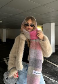 Beige nepbontjas, kleurrijke geruite sjaal met tinten roze, grijs en crème, en een wit label met de tekst "Acne Studios." Ronde zonnebril.