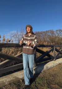 Femme portant un pull marron à motifs, un bonnet et un jean, debout sur un chemin à côté d'une clôture en bois, avec des roseaux secs et des arbres sans feuilles.