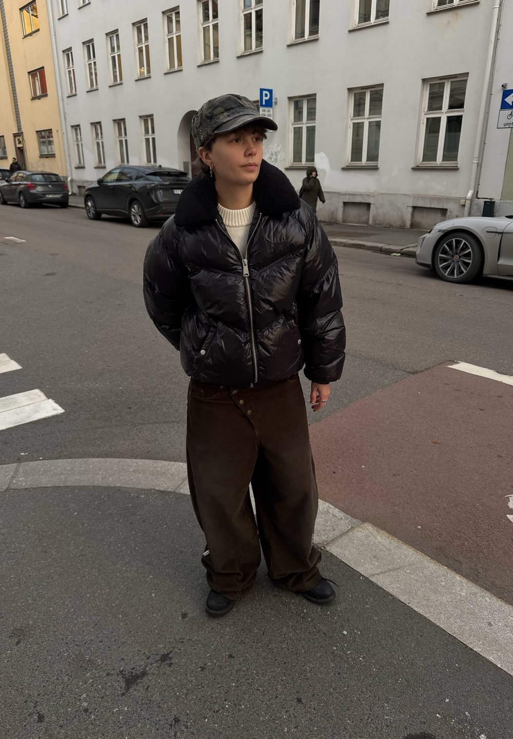 Black puffa jacket with a shearling collar, cream roll-neck jumper, wide brown trousers, and dark boots. Person stands on a city street.