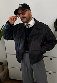 Μαύρο δερμάτινο μπουφάν μηχανής με μεγάλο γιακά και τσέπες με φερμουάρ. Συνδυασμένο με έναν μαύρο πουλόβερ και γκρίζες παντελόνες.