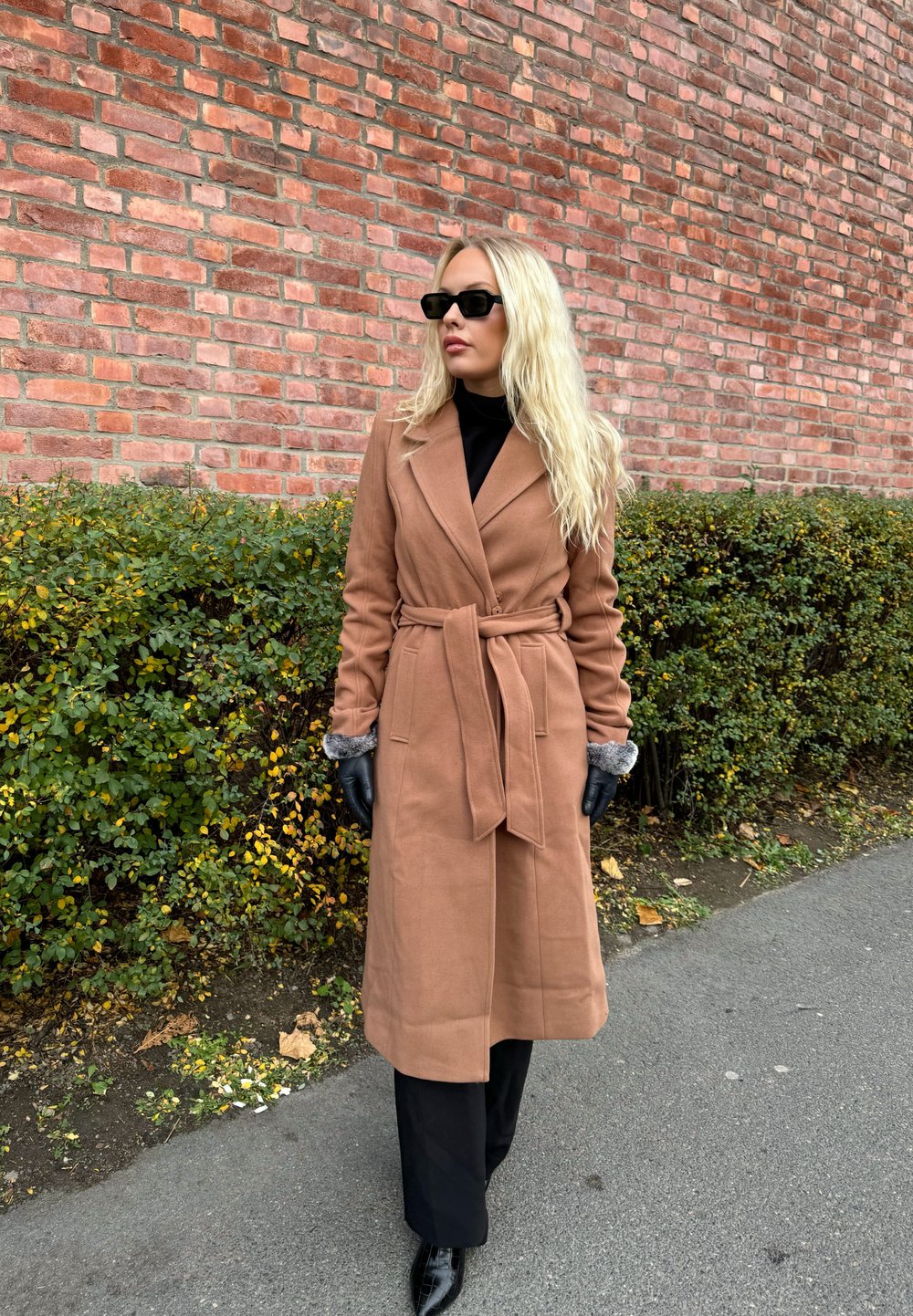 Brown wool trench coat with a belted waist, grey fuzzy cuffs, worn with black gloves and trousers, set against a brick wall and greenery.