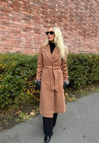 Brown wool trench coat with a belted waist, grey fuzzy cuffs, worn with black gloves and trousers, set against a brick wall and greenery.