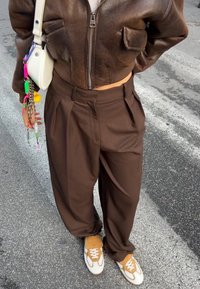 Casaco bomber cropped de couro castanho com dois bolsos frontais, combinado com calças pregueadas castanhas de cintura alta e sapatilhas brancas com detalhes em laranja.
