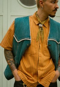 Veste en jean bleue aux bords effilochés portée par-dessus une chemise à rayures orange à boutons. Un foulard vert autour du cou ajoute du détail. Un jean foncé complète la tenue.