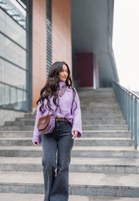 Pull en tricot violet avec un col montant, associé à un jean noir à jambes larges. Sac tressé marron avec des clous, marchant sur des escaliers en pierre.