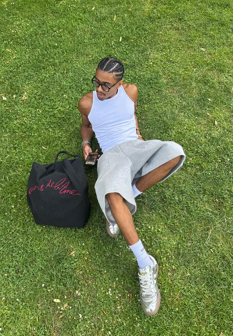 Homme aux cheveux tressés et lunettes, allongé sur l'herbe, portant un débardeur blanc, un short gris, des chaussettes blanches, des chaussures argentées, tenant un téléphone à côté d'un sac noir.