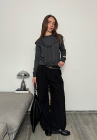 Jeune femme portant un pull gris foncé et un pantalon large noir, tenant un sac fourre-tout noir dans une pièce minimaliste avec un canapé gris et une lampe.